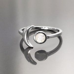 Peut inclure: Une bague argentée représentant un croissant de lune et un élément circulaire. L'élément circulaire contient une pierre blanche irisée. La bague est posée sur une surface réfléchissante, créant un effet miroir. Le design de la bague est minimaliste et céleste.