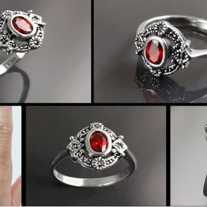 K&ouml;nnte beinhalten: Ein silberner Ring mit einem ovalen, roten Edelstein in der Mitte, umgeben von einem dekorativen floralen Design. Der Ring wird aus verschiedenen Blickwinkeln gezeigt, um seine komplizierten Details hervorzuheben. Auf einem Foto wird der Ring an einer Hand getragen.