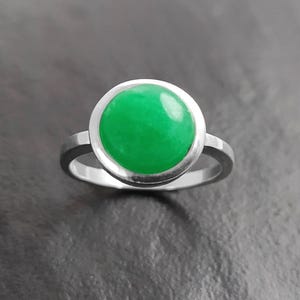 May include: A silver ring featuring a large, round, bright green gemstone. The gemstone is set in a bezel setting, and the band is a simple, polished silver. The ring is displayed against a dark gray background.