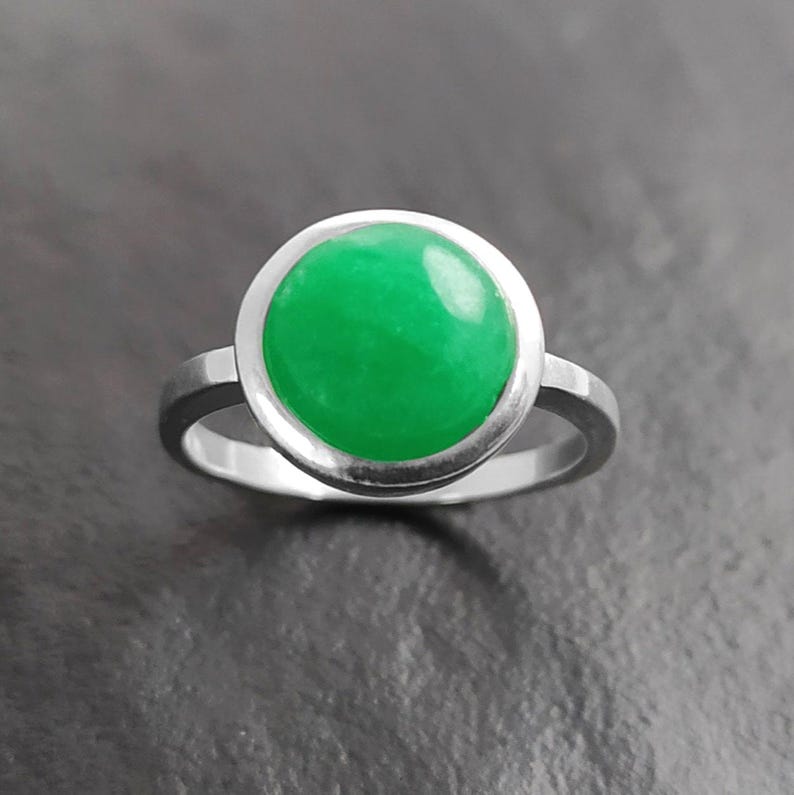 May include: A silver ring featuring a large, round, bright green gemstone. The gemstone is set in a bezel setting, and the band is a simple, polished silver. The ring is displayed against a dark gray background.