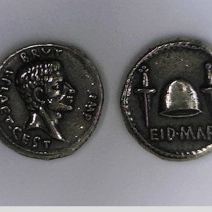 May include: Silver Roman coin with a portrait of a man on one side and a helmet with two swords on the other side. The inscription on the coin reads "BRUT. IMP. CAESAR. CEST." and "EID. MAR."