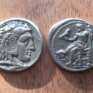 May include: Silver coin featuring a portrait of a man with long hair on one side and a seated figure holding a bird on the other. The coin has a beaded border.