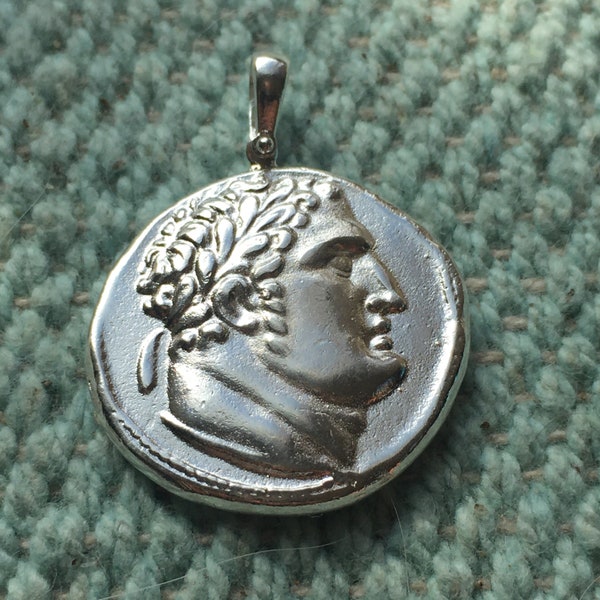 May include: Silver pendant featuring a profile of a person wearing a laurel wreath. The pendant is round and has a detailed design.