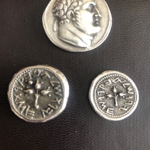 May include: Three silver coins featuring a tree design and text in an ancient language. The coins are displayed on a black leather surface.
