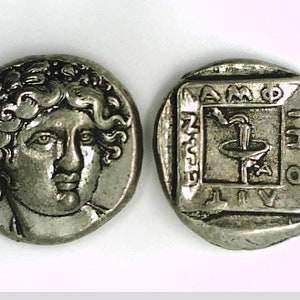 Puede incluir: Moneda de plata con el retrato de un hombre con una corona de laurel en la cabeza en un lado y una fuente estilizada con la inscripción "ZAMPHIO" en el otro lado.