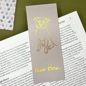 May include: A gold foil bookmark with a gray background. The bookmark features a line drawing of a dog with the text "Paws Here..." printed below the dog.