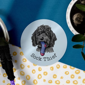 May include: A round sticker with a light blue background features a black and white illustration of a dog with a blue sock in its mouth. The text "Sock Thief" is printed in black below the dog.