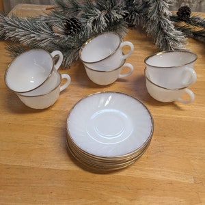 May include: Set of six white milk glass teacups with gold trim. The cups are stacked in pairs and are arranged on a wooden surface.
