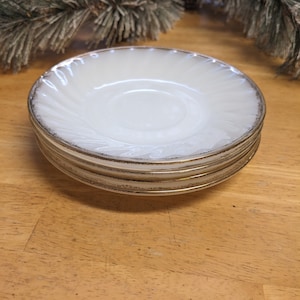 May include: A stack of four white ceramic saucers with a gold rim. The saucers have a swirl design.