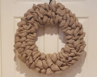 May include: A circular wreath made of natural burlap fabric. The wreath is a light brown color and has a textured, ruffled appearance. It is hanging on a white door.