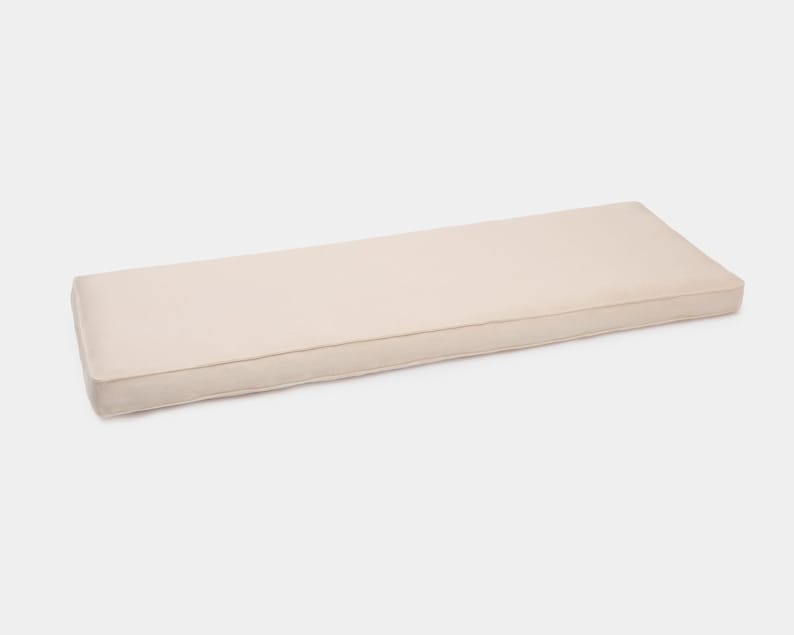 May include: A rectangular bench cushion in a solid light beige color. The cushion features a smooth, flat top surface with defined, stitched edges that create a clean, structured profile. The fabric has a uniform, matte texture throughout.