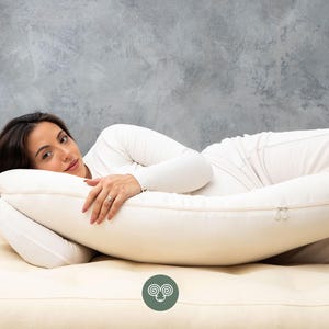 May include: A cream-colored, full-body pillow with a zipper closure. The pillow is designed for comfort and support. The person in the image is wearing a white long-sleeve shirt and white pants.