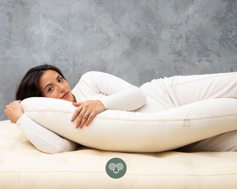 May include: A cream-colored, full-body pillow with a zipper closure. The pillow is designed for comfort and support. The person in the image is wearing a white long-sleeve shirt and white pants.