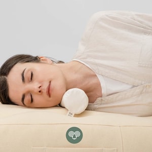 May include: A small, cylindrical, off-white neck pillow positioned under the head of a person resting on a matching off-white mattress. The pillow features a small fabric tag with a circular logo depicting a stylized ram head in white on green.