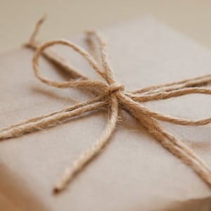 May include: A square-shaped gift box wrapped in plain, light brown kraft paper. The package is secured with a thin, natural-coloured jute twine tied into a simple bow on the top centre, creating a textured and rustic appearance for the parcel.