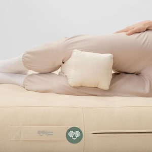 May include: A rectangular off-white knee pillow positioned between the legs of a person wearing beige pants. The pillow is made of a plain fabric and rests on a matching off-white mattress featuring a visible zipper and a Home of Wool label.