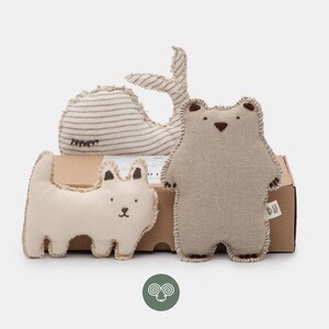May include: Three stuffed animal toys including a beige bear, a white cat, and a striped whale, all featuring visible whip-stitched edges and embroidered facial details, positioned in front of a rectangular cardboard shipping box.