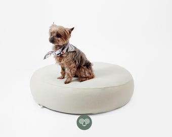 May include: A small brown and tan Yorkshire Terrier dog wearing a white bandana with a black and red abstract pattern, sitting on a large, round, off-white pet bed with a smooth, solid-coloured fabric surface and a low profile.