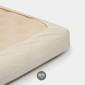 Könnte beinhalten: Ein cremefarbenes Spannbettlaken, das eine Matratzenecke bedeckt. Der Stoff hat eine glatte, schlichte Webstruktur und das Laken ist straff über die Matratzenkante gezogen. Ein kleines kreisförmiges Logo mit einem stilisierten Widderkopf ist darunter zu sehen.