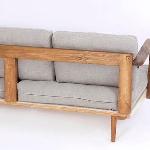 May include: A sofa featuring a natural light brown wooden frame with a square logo detail on the side. The furniture includes thick, light gray fabric cushions for the back and seat, supported by tapered wooden legs and curved armrests.