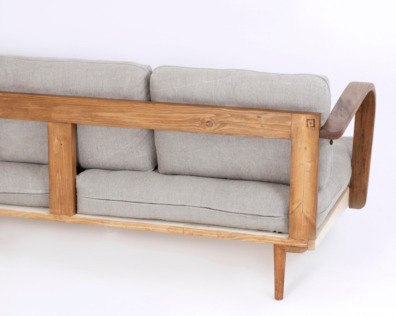 May include: A sofa featuring a natural light brown wooden frame with a square logo detail on the side. The furniture includes thick, light gray fabric cushions for the back and seat, supported by tapered wooden legs and curved armrests.