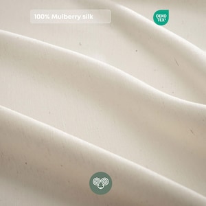 May include: A piece of off-white fabric with a smooth, draped texture. The text 100% Mulberry silk is visible in a rectangular box, alongside a green OEKO-TEX label and a circular green icon featuring a stylized ram head illustration.