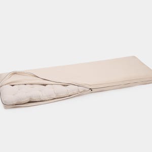 May include: A rectangular floor cushion featuring a smooth, light beige zippered cover partially opened to reveal a tufted, off-white inner mattress. The cover is made of a solid-colored fabric, while the interior shows a quilted texture.