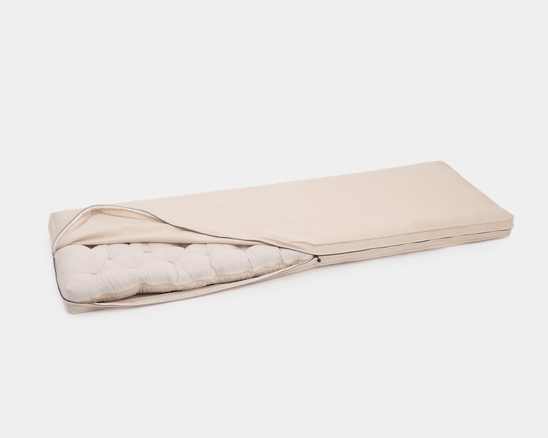 May include: A rectangular floor cushion featuring a smooth, light beige zippered cover partially opened to reveal a tufted, off-white inner mattress. The cover is made of a solid-colored fabric, while the interior shows a quilted texture.