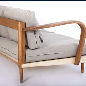 May include: A sofa featuring a natural wood frame with curved armrests and light gray linen cushions. The wooden structure displays visible grain and joinery details, supporting a seat and backrest with piped edges and a small metal zipper pull.