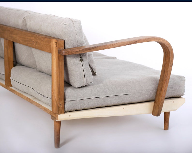 May include: A sofa featuring a natural wood frame with curved armrests and light gray linen cushions. The wooden structure displays visible grain and joinery details, supporting a seat and backrest with piped edges and a small metal zipper pull.