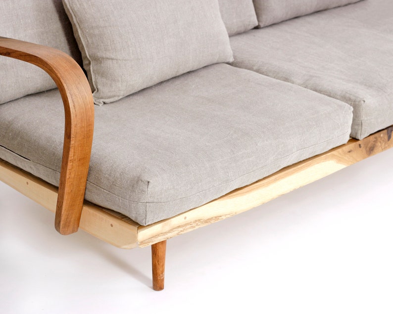 May include: A sofa featuring a light beige fabric upholstery with matching cushions, supported by a natural light-colored wooden frame with curved armrests and tapered legs. The base of the frame displays a raw, live-edge wood texture.