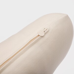 May include: Close-up of a cream-colored, cylindrical pillow with a zipper closure. The zipper is beige and runs along the side of the pillow. The pillow is made of a soft, textured fabric.