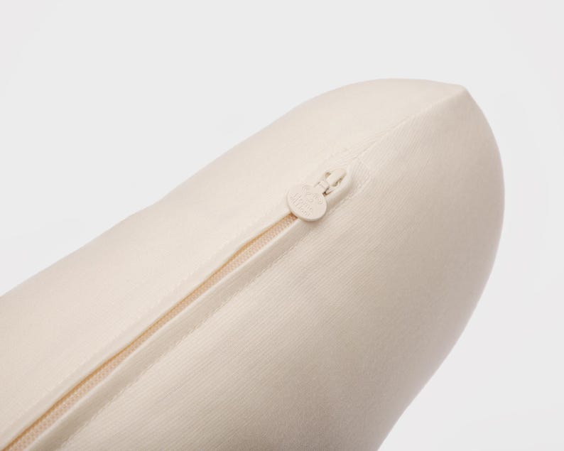 May include: Close-up of a cream-colored, cylindrical pillow with a zipper closure. The zipper is beige and runs along the side of the pillow. The pillow is made of a soft, textured fabric.