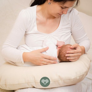 May include: A crescent-shaped nursing pillow in a solid off-white color. The pillow features a circular green logo patch centered on the front, displaying a white line-art illustration of a ram's head with curled horns.