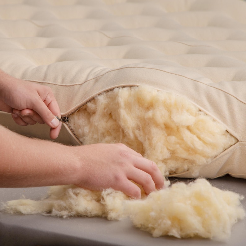 May include: A close-up of a natural-colored mattress topper with a zipper detail. The zipper is partially open, revealing the fluffy, light-yellow filling. The filling is being handled by a person.
