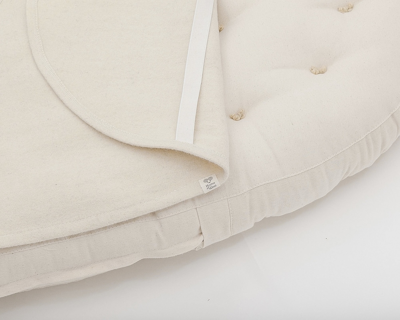 May include: A white round cushion with a removable cover. The cushion has a white fabric cover with a zipper closure and a white label with the text "The Little Green"