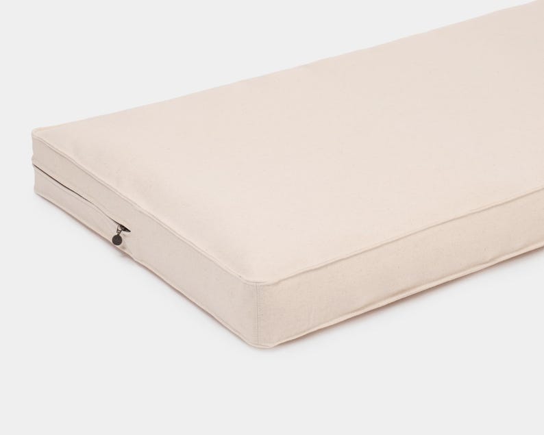 May include: A rectangular, off-white cushion featuring a smooth, solid-colored fabric cover with piped edges. A small, dark metal zipper pull is visible on the side, indicating a removable cover design for this firm, flat-profile furniture pad.