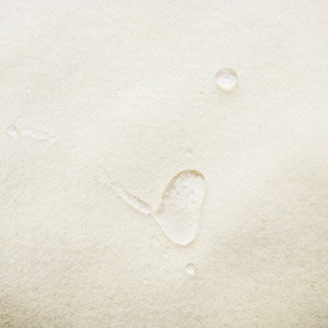 May include: A close-up of a white fabric surface with water droplets. The droplets are round and clear, and they are scattered across the surface.