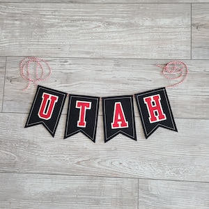 May include: A black banner with the word "UTAH" in red letters with white outlines. The banner is strung on a red and white twine. The background is a light gray wood grain.