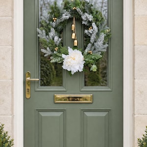 May include: A festive wreath featuring frosted green pine branches, small gold berries, and a large white peony flower. Three hanging gold bells are suspended from the centre. The wreath is mounted on a sage green door with a gold handle and letter box.
