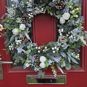 May include: A circular wreath featuring white roses, brown pinecones, and frosted green foliage. The arrangement includes small white berries and various evergreen sprigs, creating a dense, textured display mounted on a red door with a silver knocker.