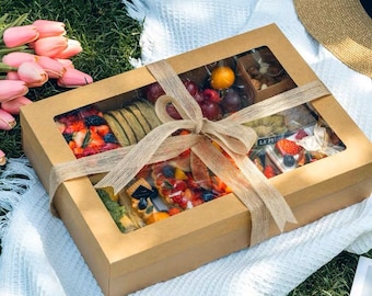 Puede incluir: Una caja de cartón marrón con una ventana transparente, llena de una variedad de bocadillos y golosinas, incluyendo fruta, pasteles y nueces. La caja está atada con una cinta marrón y está sentada sobre una manta blanca.