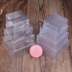 May include: A stack of clear plastic boxes, with a pink macaron in front of them. The boxes are stacked in two separate piles, one on the left and one on the right. The boxes are all the same size and shape, and they are all made of clear plastic. The macaron is a light pink color and is sitting on a wooden surface.