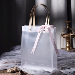 K&ouml;nnte beinhalten: Eine transparente Plastikt&uuml;te mit Holzgriffen und einer rosa Schleife aus Satinband. Die Tasche ist leer.