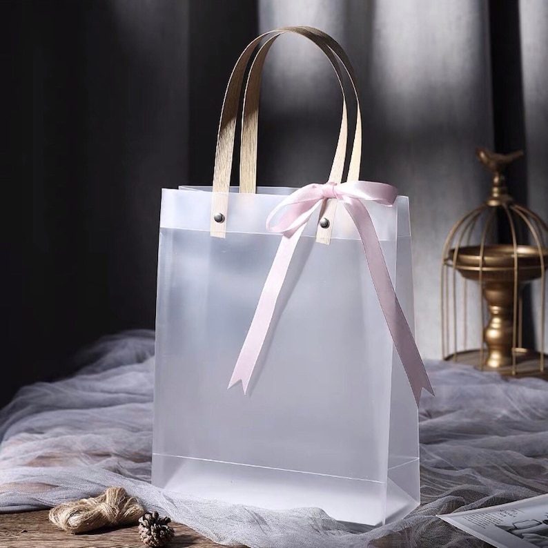 K&ouml;nnte beinhalten: Eine transparente Plastikt&uuml;te mit Holzgriffen und einer rosa Schleife aus Satinband. Die Tasche ist leer.