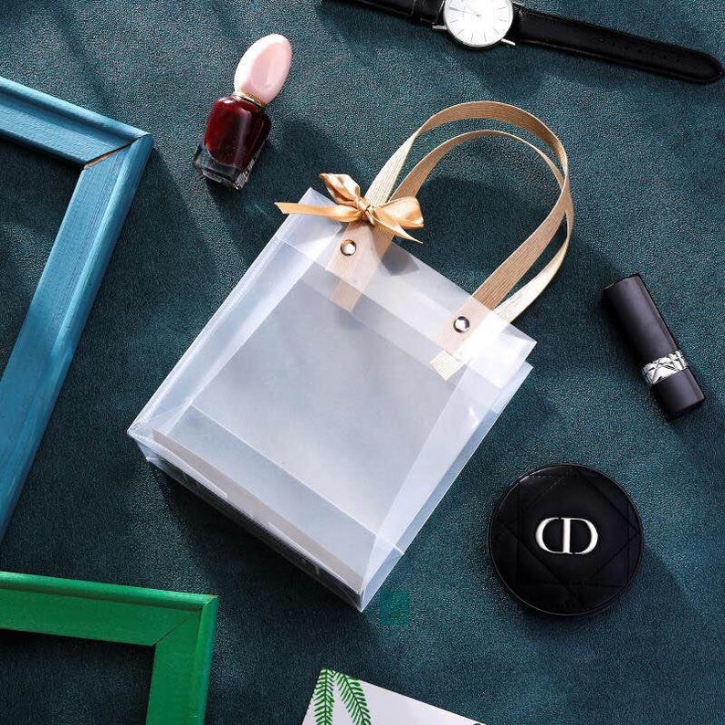 K&ouml;nnte beinhalten: Eine transparente Plastikt&uuml;te mit einer goldenen Schleife und Henkeln. Die Tasche ist leer und bereit, mit einem Geschenk gef&uuml;llt zu werden.