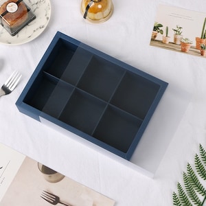 May include: A blue cardboard box with a clear plastic lid. The box has six compartments and is designed for packaging small items.