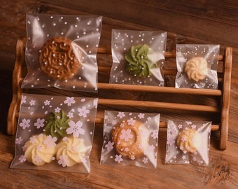 Könnte beinhalten: Transparente Plastikbeutel mit verschiedenen Mustern, darunter Punkte und rosa Blumen, sind mit verschiedenen Keksen gefüllt. Die Beutel werden auf einem Holzregal ausgestellt.