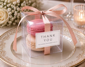 May include: A clear, square gift box tied with a pink ribbon, containing two macarons. One macaron is pink, the other is a light beige. A white label on the box reads "THANK YOU" and "FOR SHARING THIS MOMENT WITH US". The box sits on a decorative plate.
