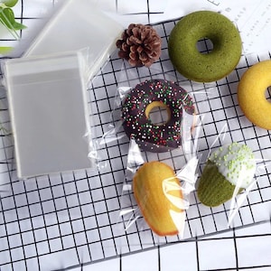 Könnte beinhalten: Transparente Plastiktüten mit einem karierten Hintergrund. Die Tüten enthalten verschiedene Backwaren, darunter ein Schokoladendonut mit Streuseln, ein grüner Teedonut, ein gelber Donut, ein Madeleines und ein grüner Tee-Kuchen mit weißem Frosting und Streuseln.